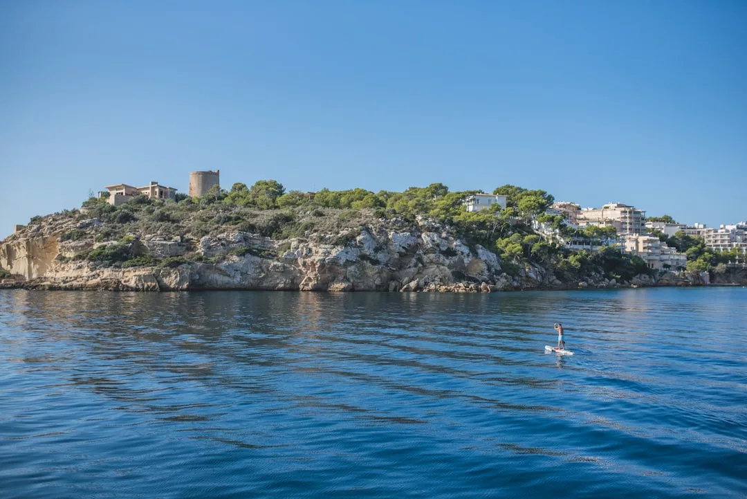 Eine Person paddelt auf einem ruhigen blauen Meer in der Nähe der felsigen Küste Mallorcas, mit Häusern, Bäumen und einem historischen Steinturm unter einem klaren Himmel.