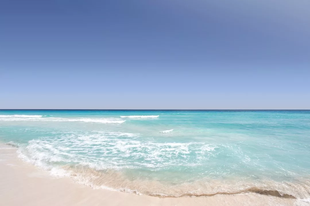 Klarer blauer Himmel über einem ruhigen, türkisfarbenen Ozean mit sanften Wellen, die an einen Sandstrand auf Mallorca schlagen.