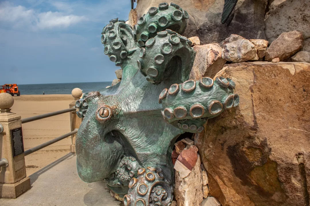 Eine große Tintenfischskulptur aus Bronze ist in eine Steinmauer am Strand auf Mallorca eingelassen, im Hintergrund sind Sand und Meer zu sehen.