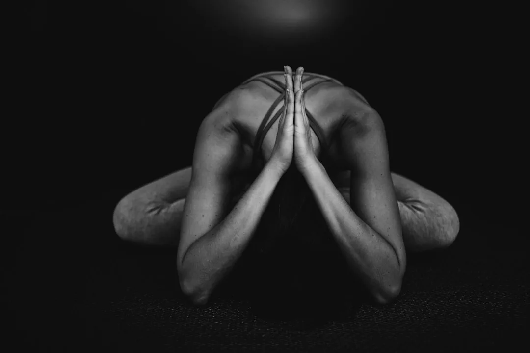 Eine Person in Sportkleidung übt eine nach vorne gebeugte Yoga-Pose mit Unterarmen und Stirn auf dem Boden, die Hände aneinandergepresst. Dieses Schwarz-Weiß-Bild fängt einen ruhigen Moment der Achtsamkeit ein, möglicherweise während eines Retreats auf Mallorca.