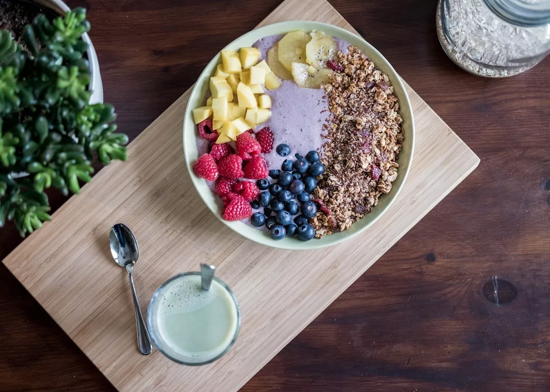 Eine Smoothie-Schüssel mit Mango, Banane, Himbeeren, Blaubeeren und Müsli steht auf einem Holzbrett neben einem Löffel und einem Glas grünem Getränk und erinnert an die frischen Aromen Mallorcas.