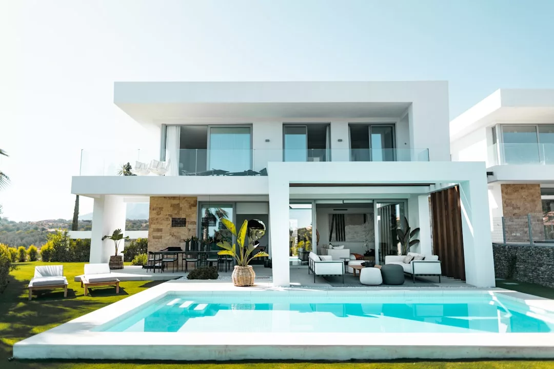 Modernes zweistöckiges Haus auf Mallorca mit großen Fenstern, einer Außenterrasse, Liegestühlen und einem Swimmingpool im Vordergrund unter klarem Himmel.