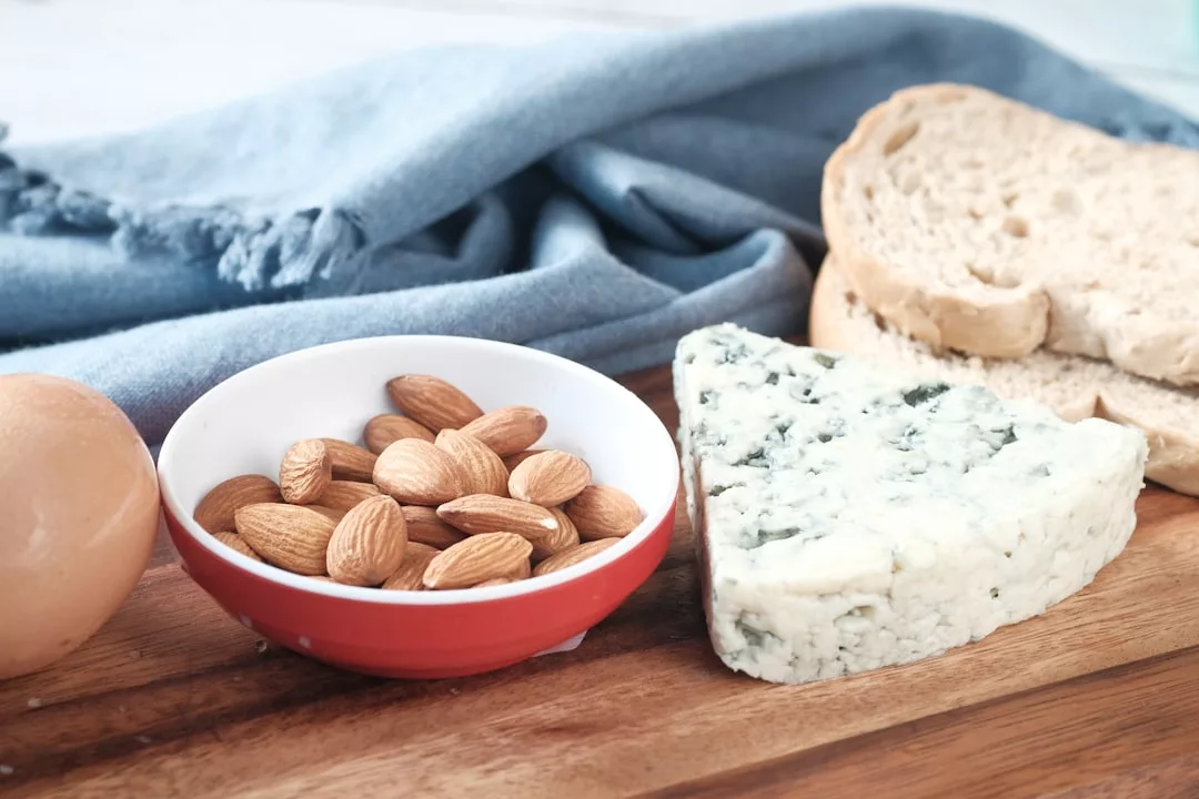 Ein Holzbrett mit einem Stück Blauschimmelkäse, einer Schale Mandeln, einem braunen Ei und Brotscheiben erinnert an die rustikalen Aromen Mallorcas, im Hintergrund ein blaues Tuch.