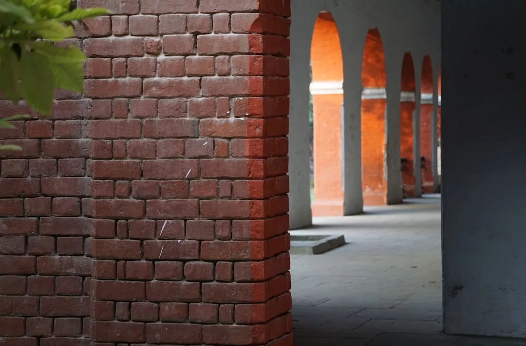Im Vordergrund steht eine rote Backsteinmauer, während sich rechts im Hintergrund ein Korridor aus roten Backsteinbögen und -säulen erstreckt, was an den architektonischen Charme erinnert, der oft auf Mallorca zu finden ist.