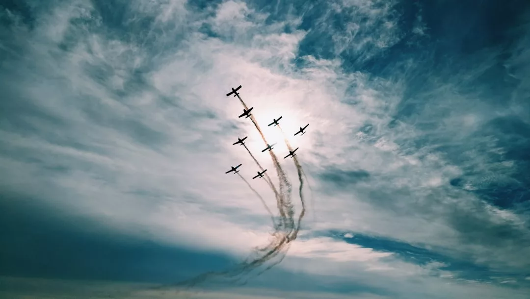 Neun Flugzeuge fliegen in Formation über Mallorca und hinterlassen Rauchfahnen vor einem teilweise bewölkten Himmel, hinter dem die Sonne teilweise verborgen ist.