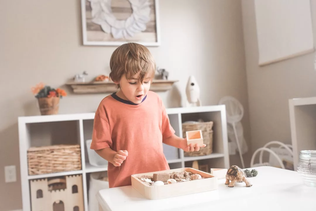 Ein kleiner Junge in einem orangefarbenen Hemd sieht überrascht aus, während er an einem weißen Tisch in einem hellen Raum mit einer hölzernen Sensorikbox spielt, was an die hellen, luftigen Innenräume erinnert, die man oft auf Mallorca findet.