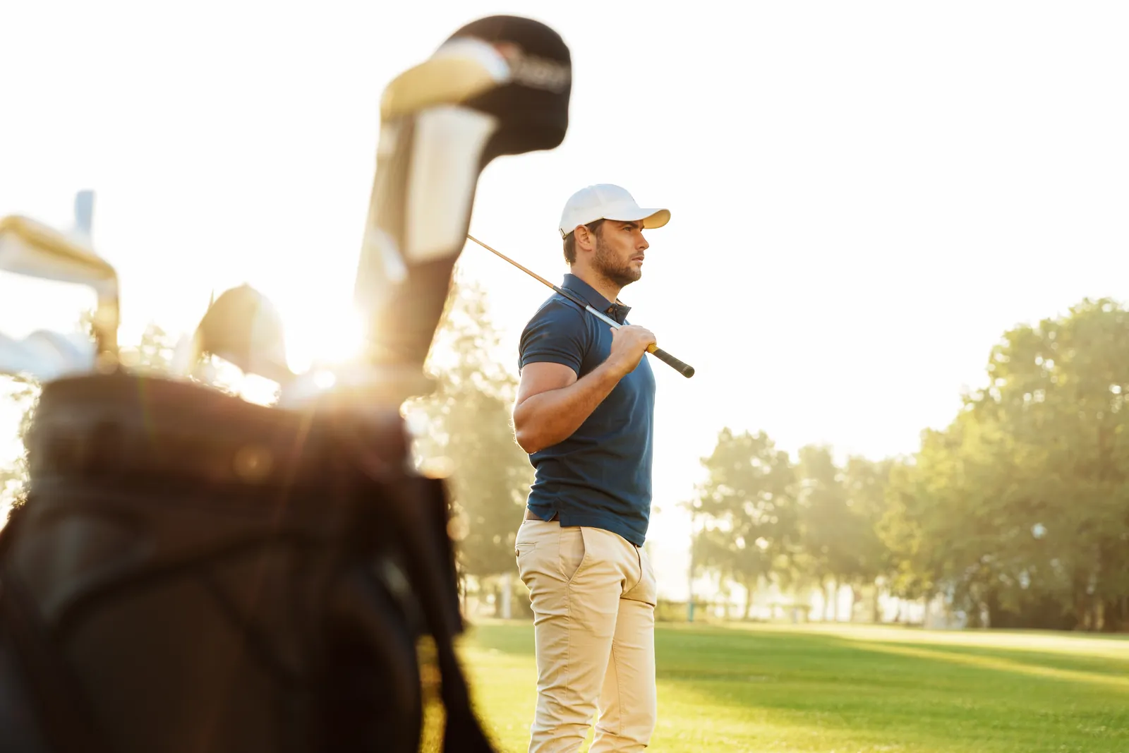 Golfspieler mit Schläger über der Schulter auf einem Fairway im Gegenlicht, im Vordergrund ein Golfbag
