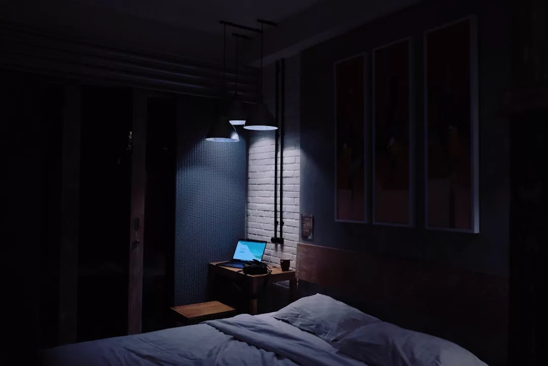 Ein schwach beleuchtetes Schlafzimmer mit einem ungemachten Bett im Vordergrund und einem Schreibtisch mit einem offenen Laptop, der im Hintergrund von zwei Hängelampen beleuchtet wird und an einen ruhigen Abend auf Mallorca erinnert.