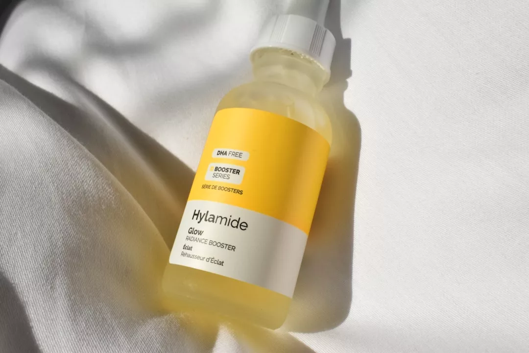 Eine Flasche Hylamide Glow Radiance Booster Serum mit gelb-weißem Etikett liegt auf einer weißen Stoffoberfläche und fängt die sonnige Essenz Mallorcas ein.