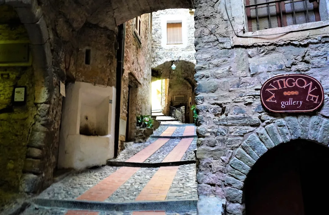 Eine schmale Steingasse mit gestreiftem Gehweg führt bergauf an rustikalen Gebäuden auf Mallorca vorbei; an der rechten Wand steht ein Schild mit der Aufschrift „Antigone Gallery“.