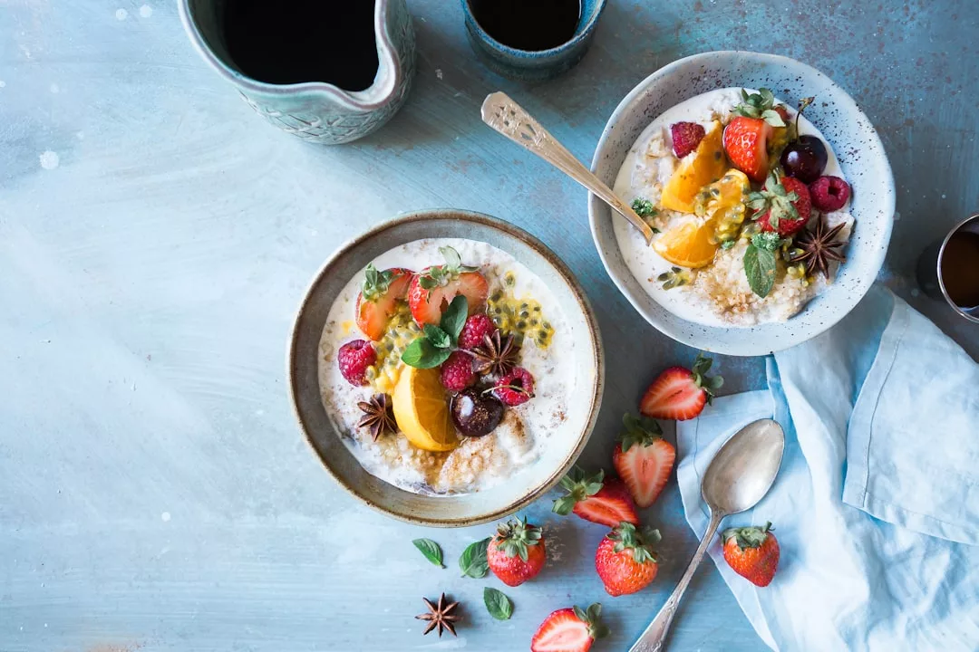 Zwei Schüsseln Joghurt, garniert mit verschiedenen frischen Früchten, Minzblättern und Sternanis, erinnern an Mallorca, begleitet von Erdbeeren, Löffeln und Tassen Kaffee auf einer hellblauen Tischplatte.