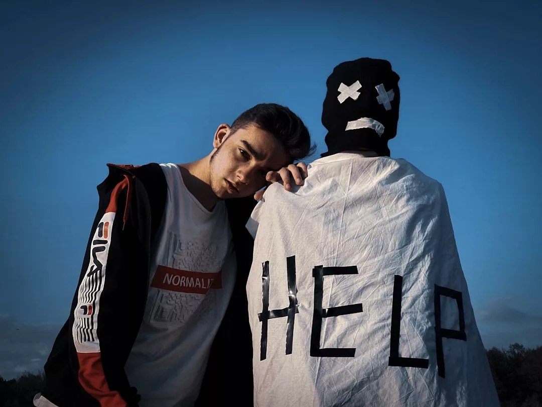 Eine Person lehnt sich an eine Gestalt mit Maske und weißem Laken, auf dessen Rücken „HELP“ steht; unter dem klaren Himmel Mallorcas erstreckt sich im Hintergrund der blaue Horizont.
