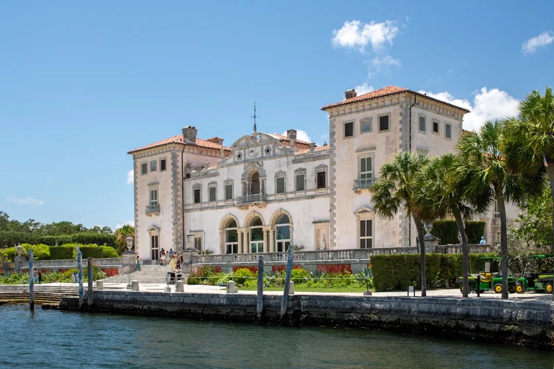 Ein großes, historisches Herrenhaus auf Mallorca mit Bogenfenstern und einem roten Ziegeldach liegt am Wasser, umgeben von gepflegten Gärten und Palmen unter einem blauen Himmel.