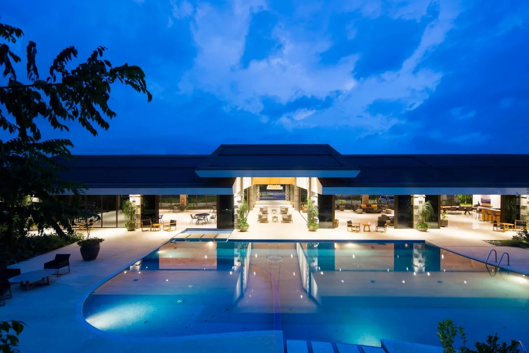 Modernes Gebäude auf Mallorca mit großen offenen Räumen und Glaswänden, das in der Abenddämmerung beleuchtet wird. Im Vordergrund befindet sich ein großer Swimmingpool, der Licht und Himmel reflektiert.