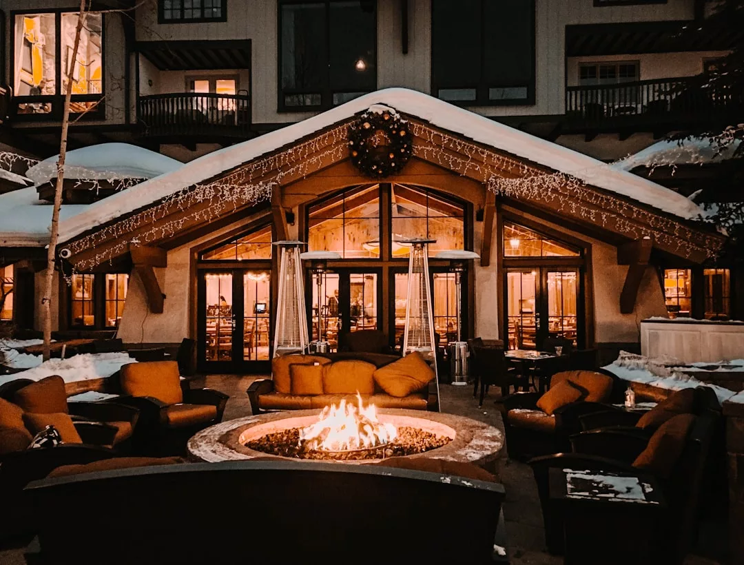 Außenterrasse einer Lodge auf Mallorca mit gepolsterten Stühlen, die um eine Feuerstelle angeordnet sind, Schnee auf dem Dach und warmes Licht, das in der Abenddämmerung im Inneren des Gebäudes leuchtet.