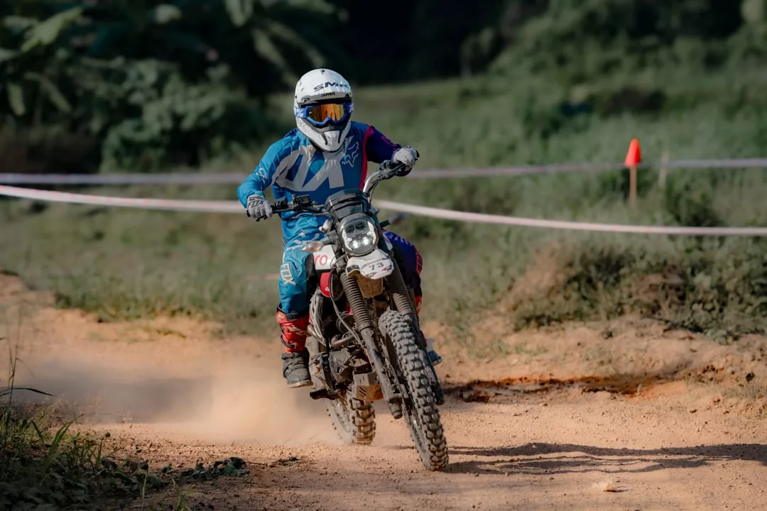 Eine Person in blauer Motocross-Ausrüstung und Helm fährt mit einem Dirtbike auf einem staubigen Offroad-Pfad auf Mallorca, im Hintergrund sind Grünflächen und ein Verkehrskegel zu sehen.