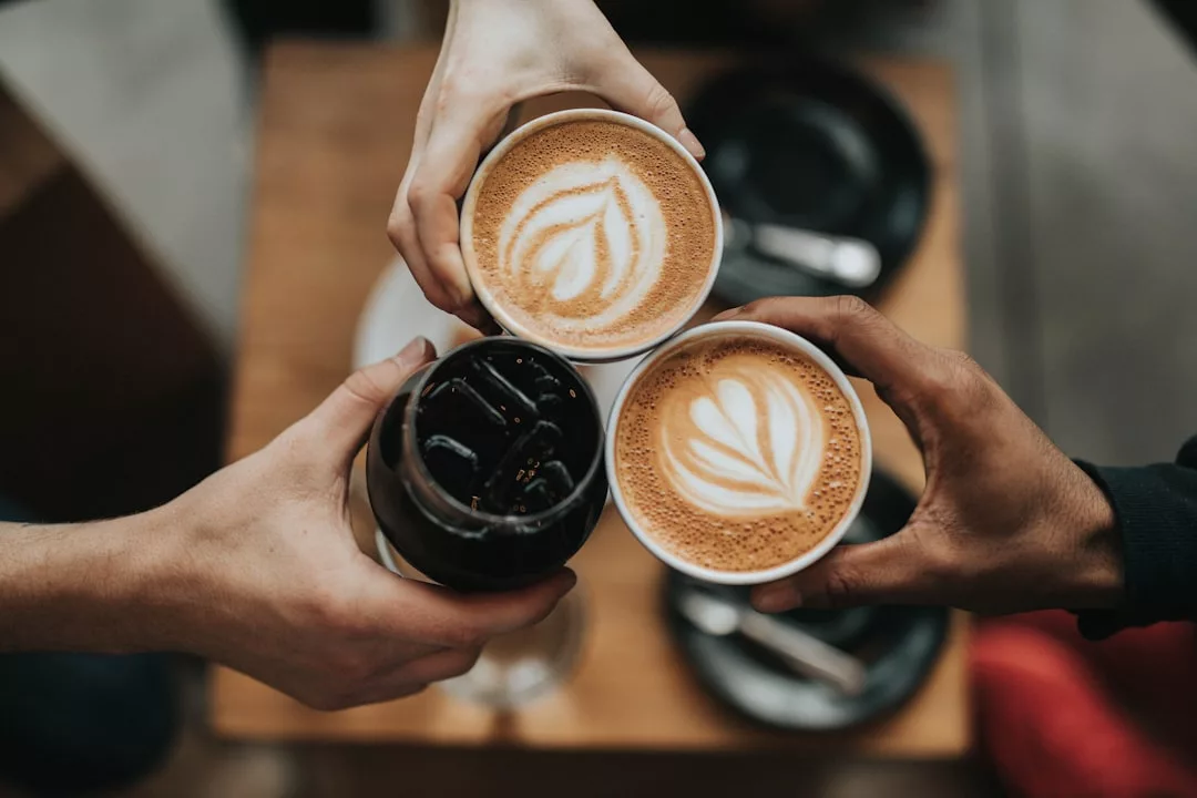 Drei Hände halten Getränke über einem Tisch auf Mallorca – zwei mit Lattes mit Latte Art und eine mit einem Glas dunkler Limonade – während darunter Teller und Besteck zu sehen sind.