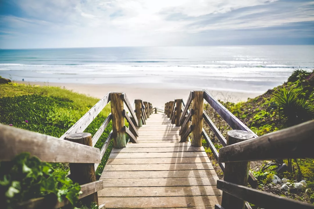 Eine Holztreppe führt hinunter zu einem Sandstrand auf Mallorca. Im Hintergrund sind Meereswellen zu sehen, der Himmel ist teilweise bewölkt. Grünes Gras und Pflanzen säumen die Seiten der Treppe.