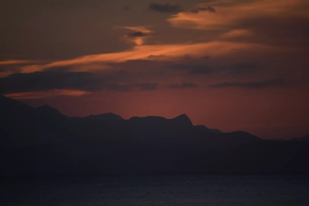 Dunkle Bergsilhouetten unter einem dramatischen orange-roten Himmel bei Sonnenuntergang, mit einem ruhigen Gewässer im Vordergrund – ein Bild, das die atemberaubende Naturschönheit Mallorcas einfängt.