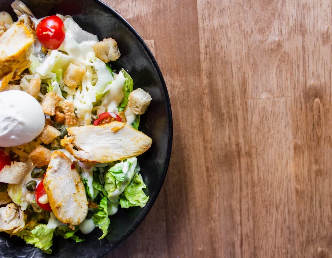 Eine Schüssel Caesar Salad mit Hühnchen, Salat, Kirschtomaten, Croutons, Kapern und einem weichgekochten Ei, serviert auf einem Holztisch auf Mallorca.