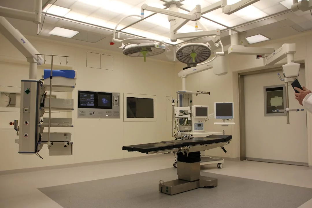 Moderner Krankenhaus-Operationssaal auf Mallorca mit Operationstisch, Deckenbeleuchtung, medizinischen Monitoren und Geräten. Auf der rechten Seite ist die Hand einer Person zu sehen, die ein Gerät hält.