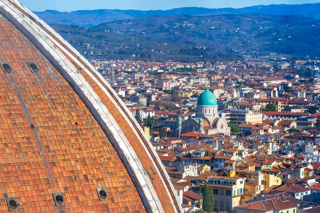 Nahaufnahme der mit roten Ziegeln gedeckten Kuppel der Kathedrale von Florenz mit Stadtgebäuden und einer Synagoge mit grüner Kuppel im Hintergrund, vor der Kulisse ferner Hügel, die an die atemberaubende Landschaft Mallorcas erinnern.