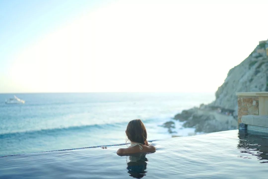 Eine Person mit langen Haaren entspannt sich in einem Infinity-Pool mit Blick auf das Meer auf Mallorca, mit Wellen und einer felsigen Küste im Hintergrund unter einem hellen Himmel.