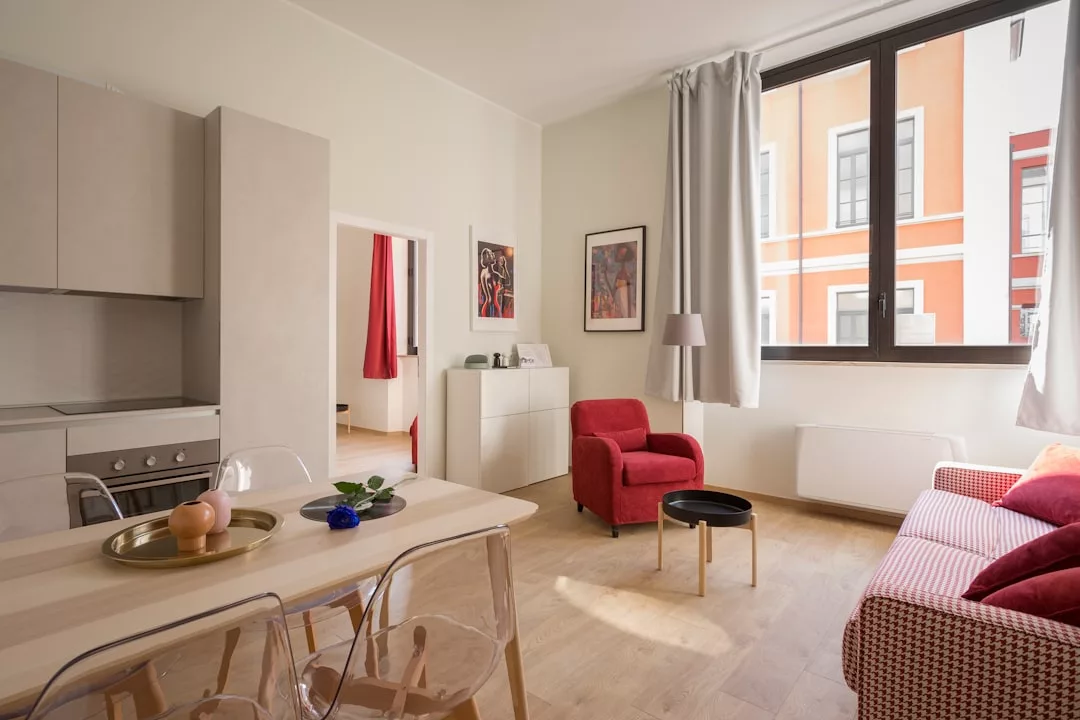 Ein modernes, sonnendurchflutetes Wohnzimmer auf Mallorca mit einem roten Sessel, einem roten Sofa, einem kleinen runden Tisch und einem Esstisch neben einer Küchenzeile; großes Fenster und Wandkunst sichtbar.