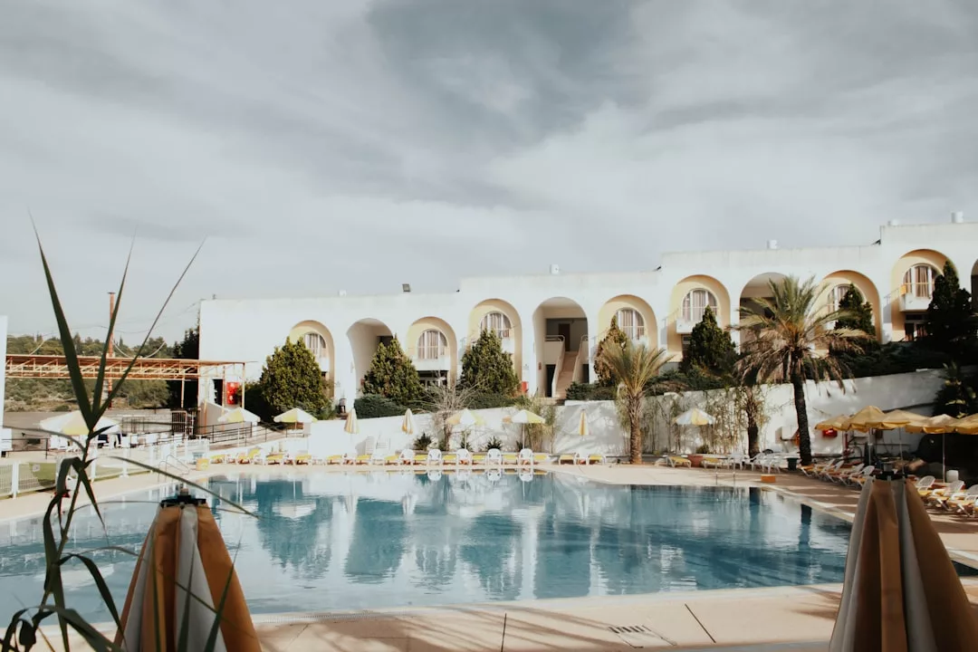 Ein großer Außenpool auf Mallorca, umgeben von Liegestühlen und Sonnenschirmen, mit einem weißen Gebäude mit Bogenfenstern und Türen im Hintergrund.