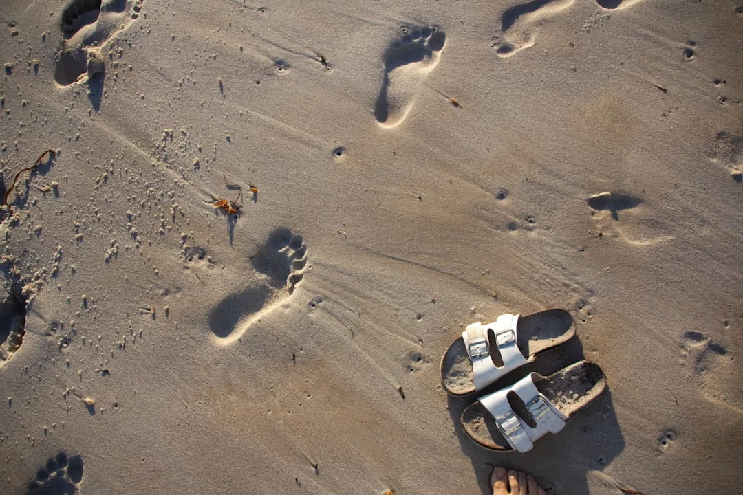 Ein Paar weiße Sandalen liegt neben mehreren menschlichen Fußabdrücken und kleinen Kieselsteinen auf einem Sandstrand auf Mallorca.