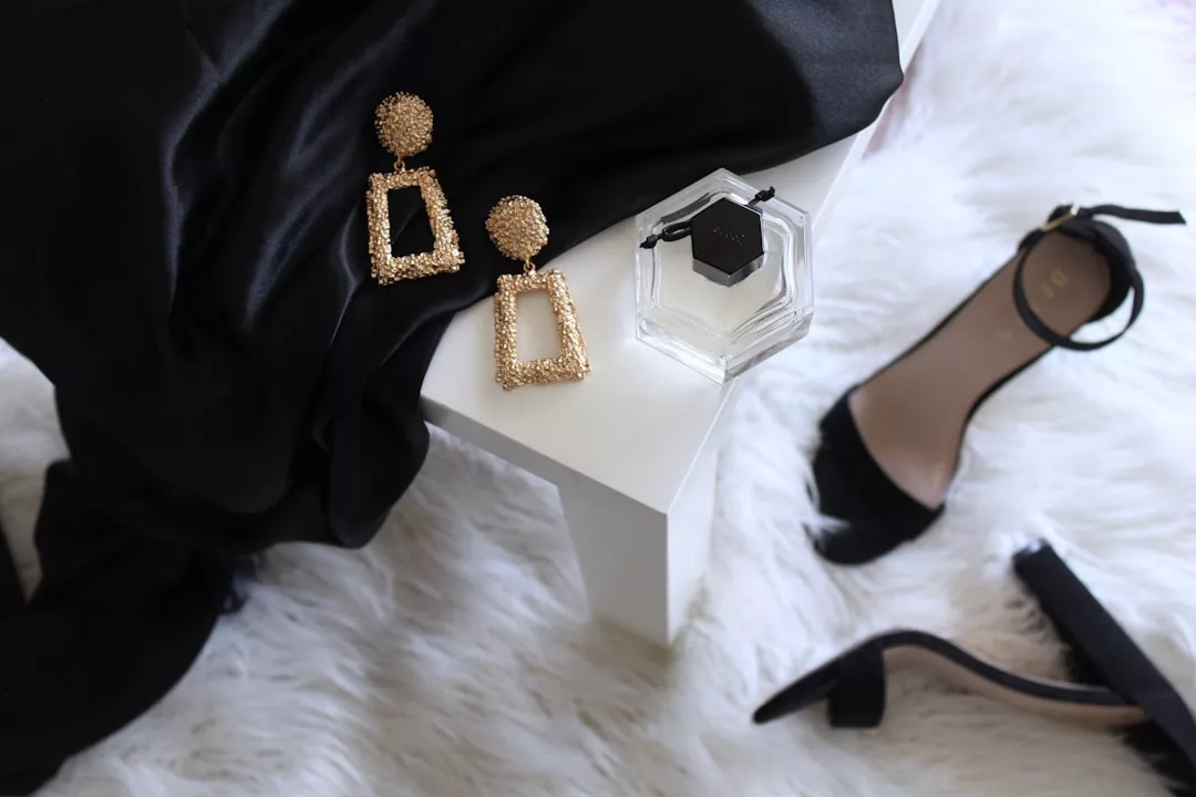 Ein Paar geometrische Ohrringe aus Gold, ein sechseckiger Parfümflakon, schwarze Sandalen mit hohen Absätzen und ein schwarzer Stoff erinnern an die schicke Eleganz Mallorcas, alles kunstvoll arrangiert auf einem weißen Tisch und einem weißen Fellteppich.