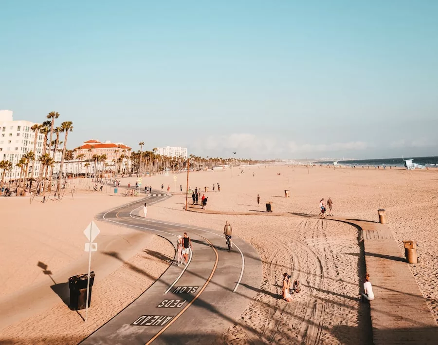 Ein breiter Sandstrand auf Mallorca mit einem asphaltierten Rad- und Fußweg, Spaziergängern und Radfahrern, Palmen und Gebäuden im Hintergrund unter einem klaren Himmel.