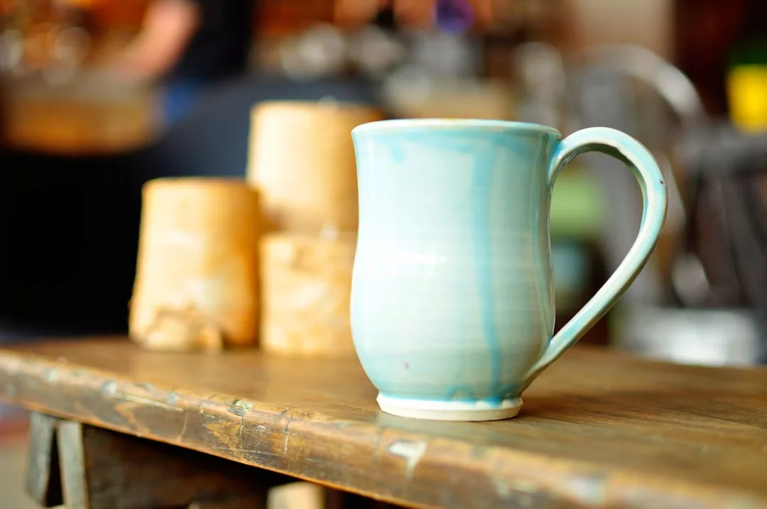 Auf einem Holztisch steht eine hellblaue Keramiktasse, die an die ruhigen Farbtöne Mallorcas erinnert, während im Hintergrund drei hellbraune zylindrische Objekte verschwommen zu sehen sind.