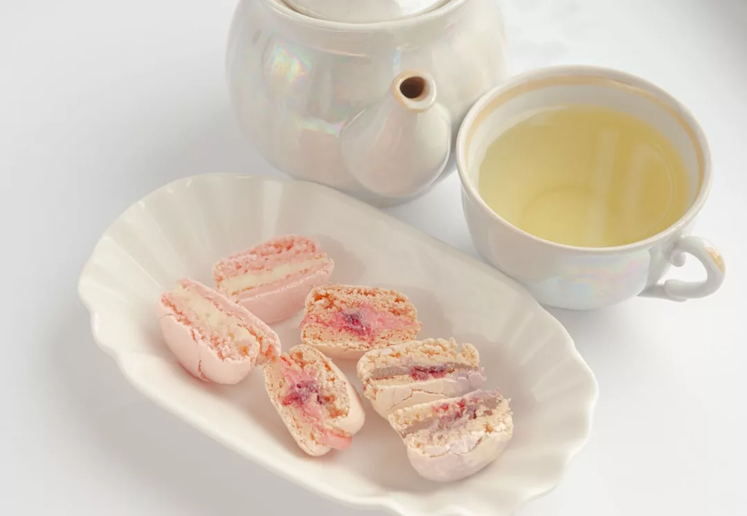 Eine weiße Teekanne, eine Tasse Tee und ein Teller mit drei halbierten rosa Macarons, gefüllt mit Sahne und roter Marmelade, sind auf einer weißen Oberfläche angeordnet und erinnern an die Eleganz eines mallorquinischen Nachmittagstees.