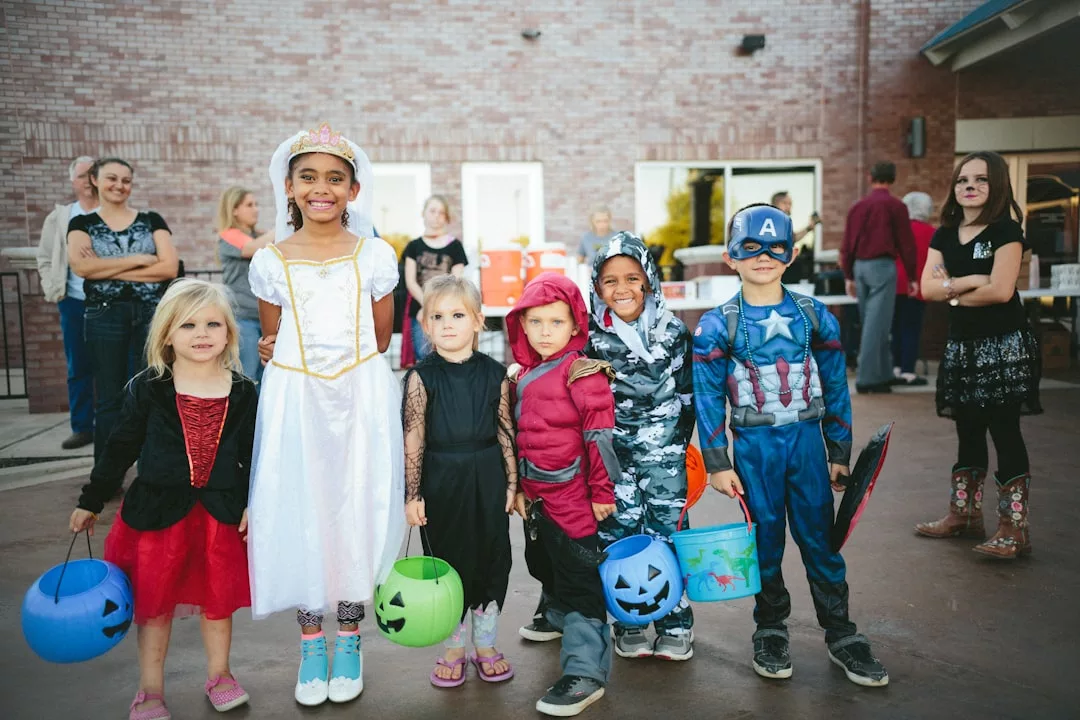 Eine Gruppe von Kindern in verschiedenen Kostümen steht im Freien auf Mallorca und hält Halloween-Eimer in der Hand. Im Hintergrund sind Erwachsene und ein Gebäude zu sehen.