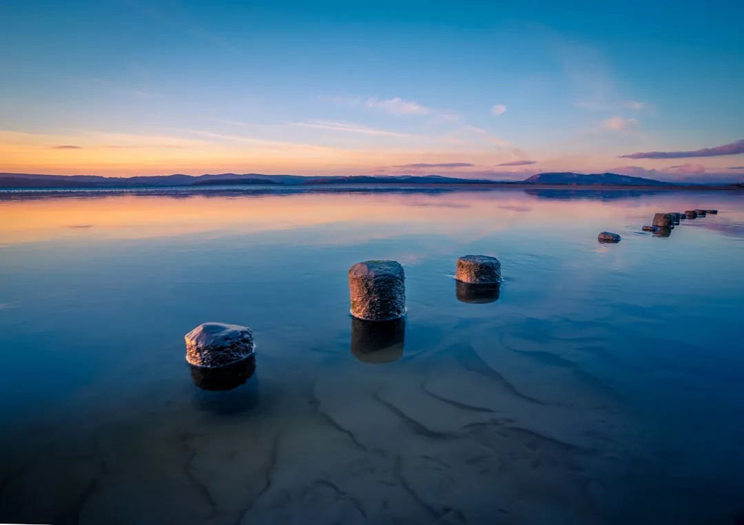 Teilweise im seichten Wasser versunkene Steinpfosten führen in die Ferne unter einem farbenfrohen Sonnenuntergangshimmel Mallorcas, am Horizont sind Berge zu sehen.