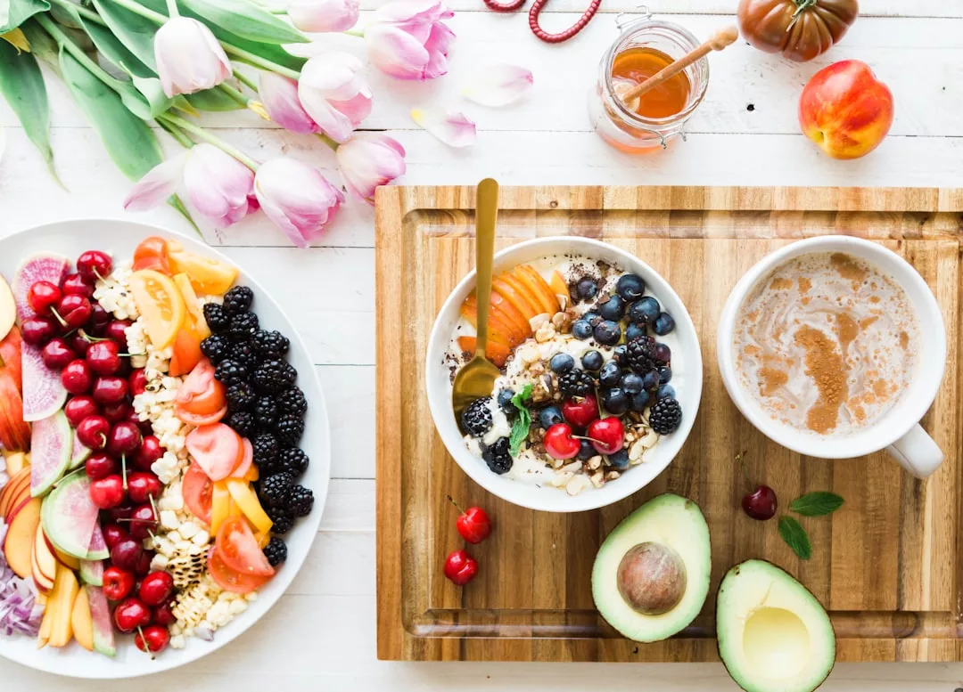 Auf einem Holztablett stehen eine Schüssel Müsli mit Früchten, eine Tasse Kaffee und eine halbierte Avocado – ein Hauch von Mallorca. Daneben steht ein Teller mit frischem Obst und einem Strauß rosa Tulpen.