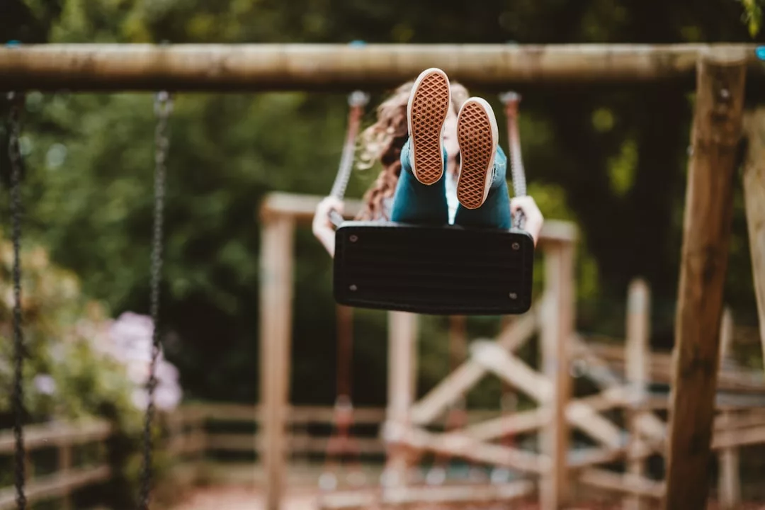 Eine Person liegt auf dem Rücken auf einer Spielplatzschaukel auf Mallorca, die Beine nach oben gestreckt, im Hintergrund sind Bäume und hölzerne Spielgeräte zu sehen.