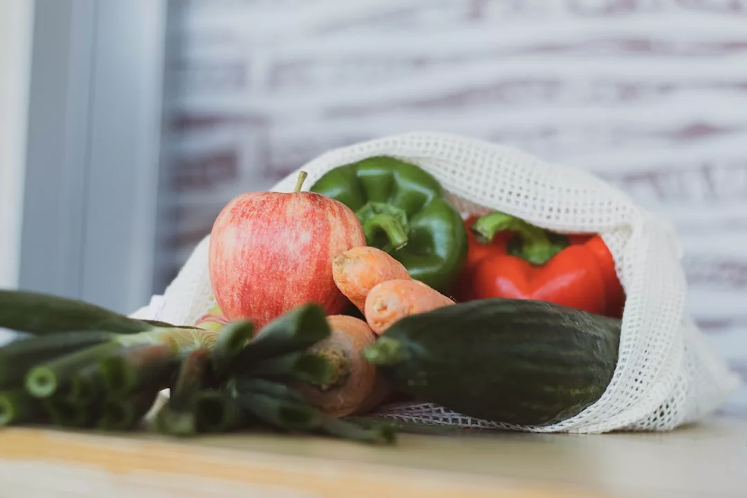 Ein wiederverwendbarer Netzbeutel gefüllt mit frischem Gemüse und einem Apfel, darunter Gurken, Karotten, Frühlingszwiebeln, eine grüne Paprika und eine rote Paprika, liegt auf einer Holzoberfläche – perfekt für den Markteinkauf auf Mallorca.
