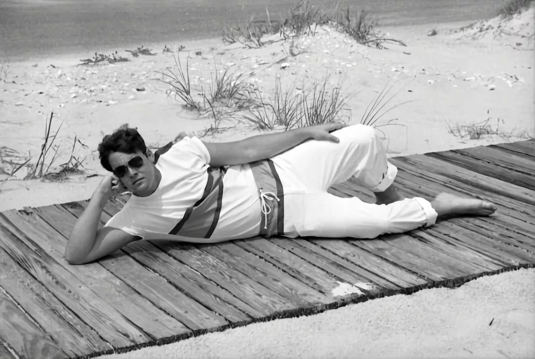 Ein Mann mit Sonnenbrille und weißer Kleidung liegt seitlich auf einem Holzsteg am Strand auf Mallorca, im Hintergrund Sand und spärliche Vegetation.