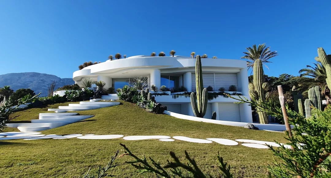 Modernes weißes Haus mit geschwungener Architektur auf Mallorca, umgeben von Kakteen, Palmen und Steinstufen auf einem grasbewachsenen Hügel unter einem klaren blauen Himmel.
