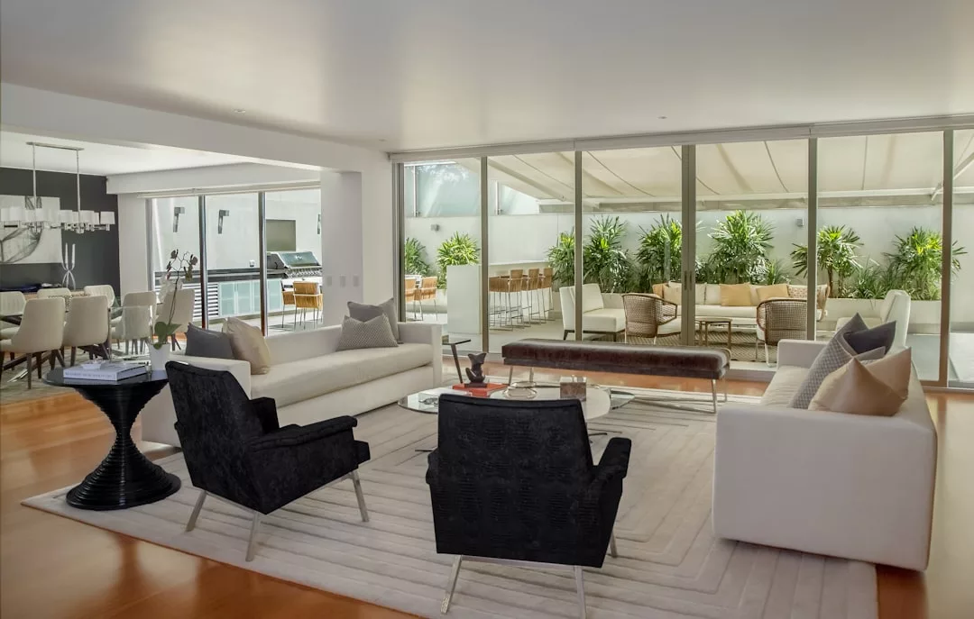 Modernes Wohnzimmer mit weißen Sofas, schwarzen Sesseln und einem Teppich, angrenzend an einen Essbereich und eine Terrasse mit Pflanzen, getrennt durch raumhohe Glastüren, die die entspannte Eleganz des Mallorca-Stils einfangen.