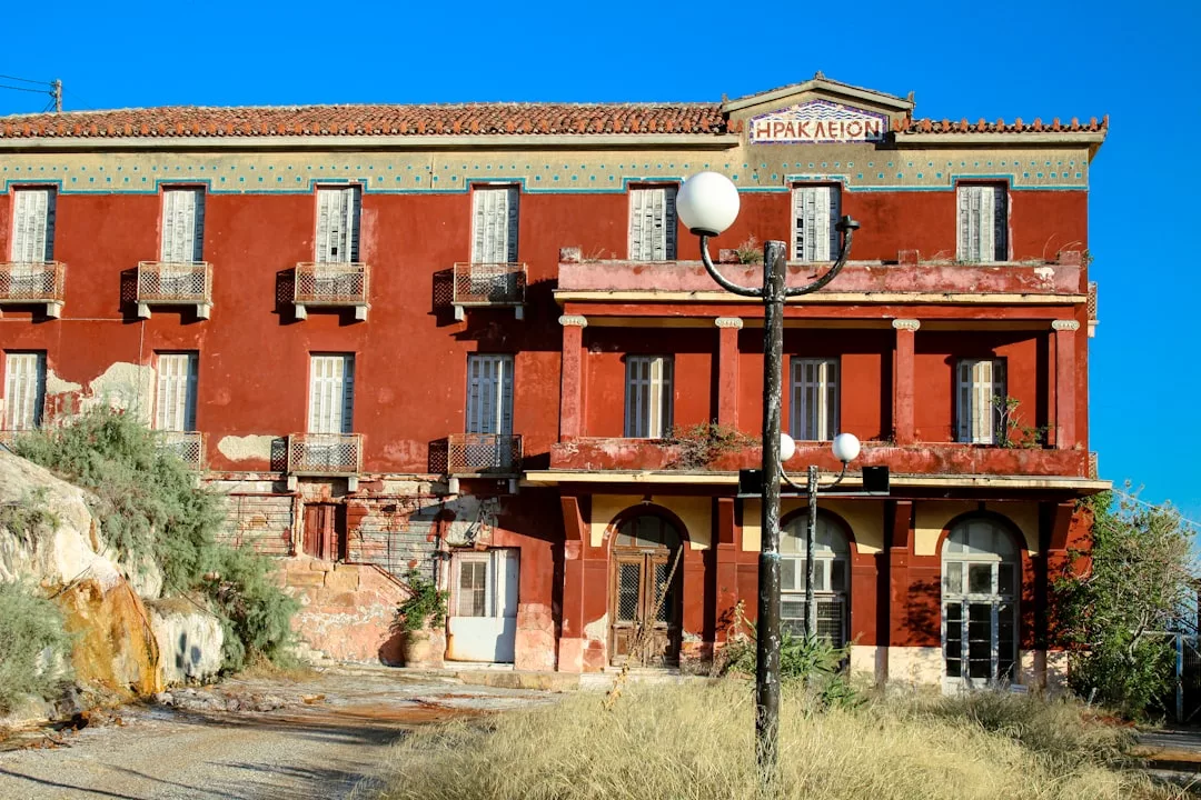 Ein großes, verwittertes rotes Gebäude mit Balkonen und einem Schild mit der Aufschrift „ΗΡΑΚΛΕΙΟΝ“ steht unter einem klaren blauen Himmel, umgeben von trockenem Gras und einem schwarzen Laternenpfahl davor und erinnert an den rustikalen Charme Mallorcas.