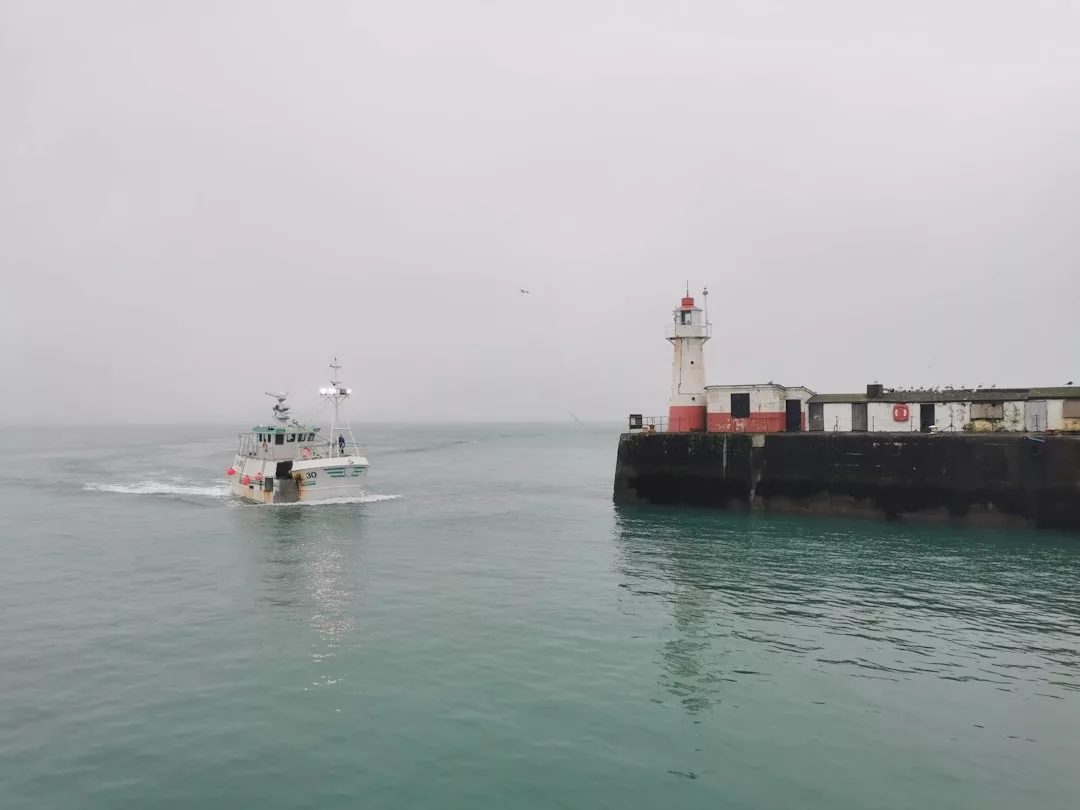 An einem bewölkten, bedeckten Tag auf Mallorca entfernt sich ein Fischerboot von einem Pier mit einem weiß-roten Leuchtturm.