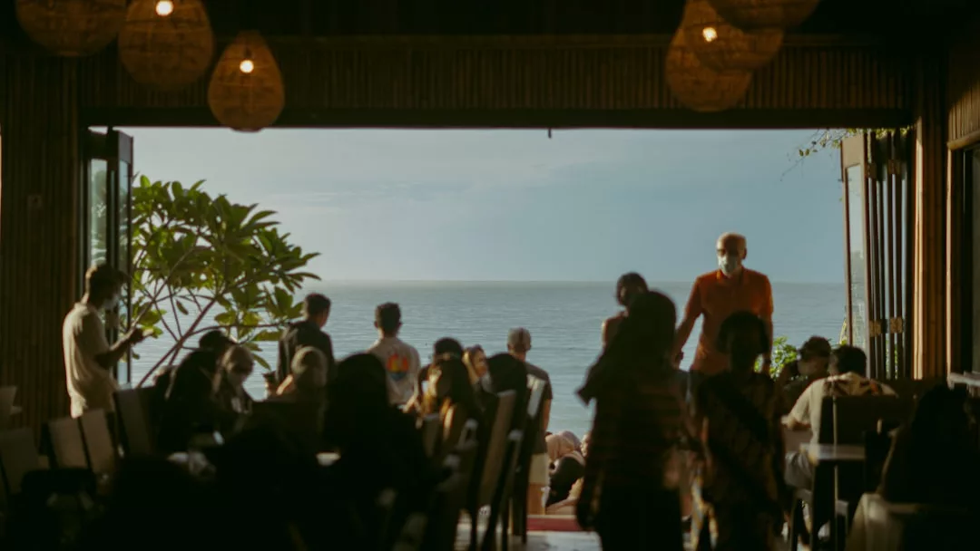 Menschen versammeln sich in einem schwach beleuchteten Restaurant auf Mallorca. Die offenen Türen blicken auf das Meer, und im Hintergrund erhellt das Sonnenlicht das Meer und den Himmel.