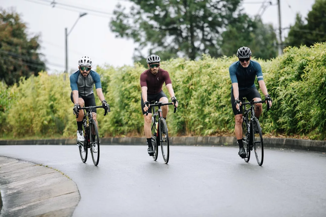 Drei Radfahrer mit Helmen und Radsportbekleidung fahren mit Rennrädern auf einer kurvigen, nassen Straße auf Mallorca, die von üppigen grünen Büschen und Bäumen gesäumt ist.