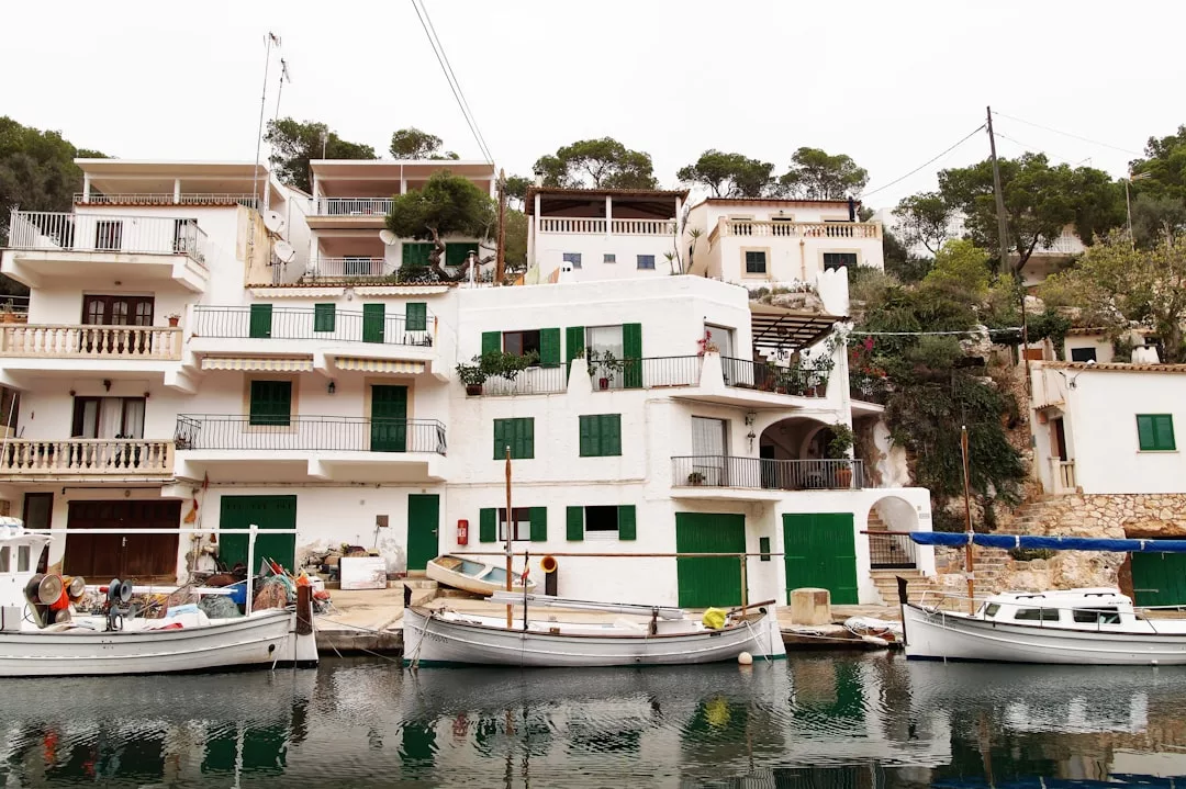 Weiße Häuser mit grünen Fensterläden und Balkonen säumen eine Uferpromenade auf Mallorca, vor der mehrere kleine Boote ankern; im Hintergrund sind Bäume und felsiges Gelände zu sehen.