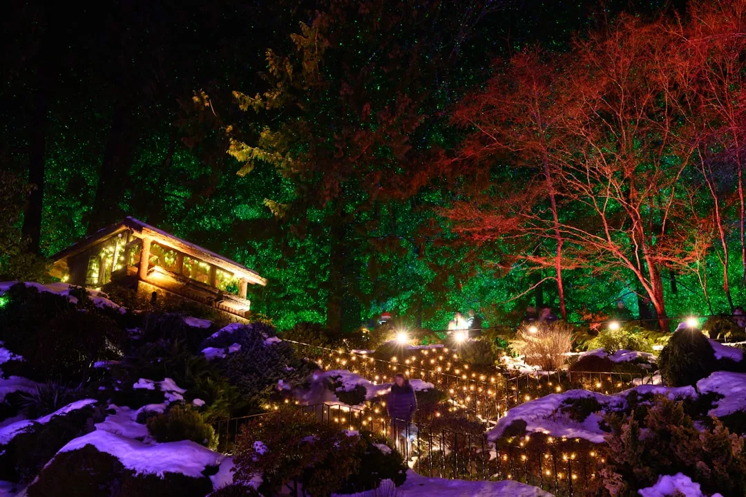 Ein verschneiter Garten wird nachts mit Lichterketten, einem kleinen Haus und bunter Beleuchtung an den Bäumen beleuchtet und ruft eine festliche Winterlandschaft hervor, die an die bezaubernden Weihnachtsdekorationen Mallorcas erinnert.
