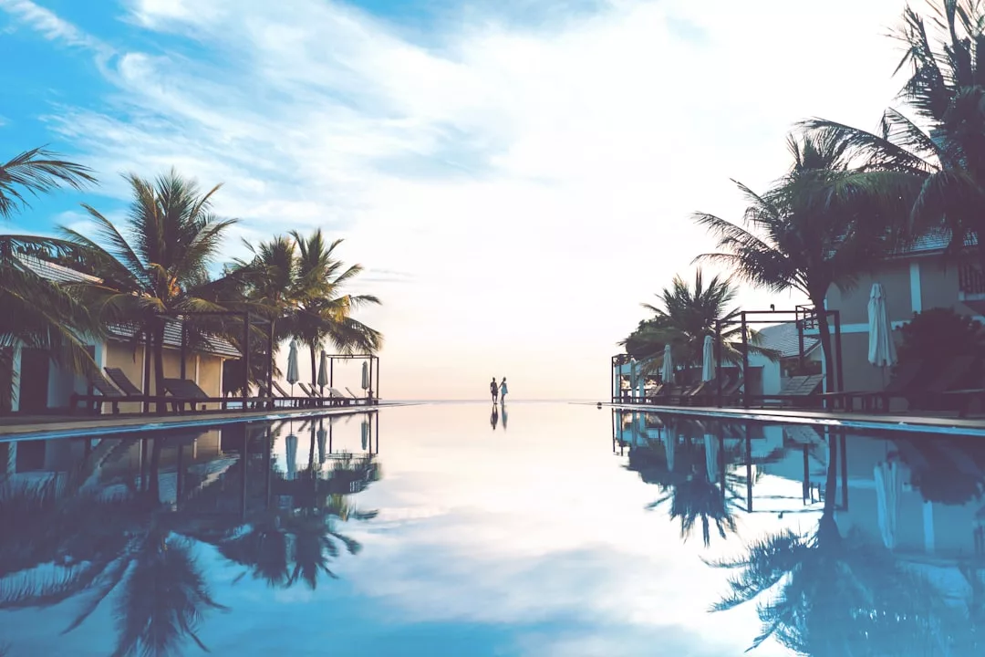 Infinity-Pool auf Mallorca, eingerahmt von Palmen, Liegestühlen und charmanten Poolgebäuden, mit zwei Personen, die am Rand vor einem strahlenden Himmel stehen.