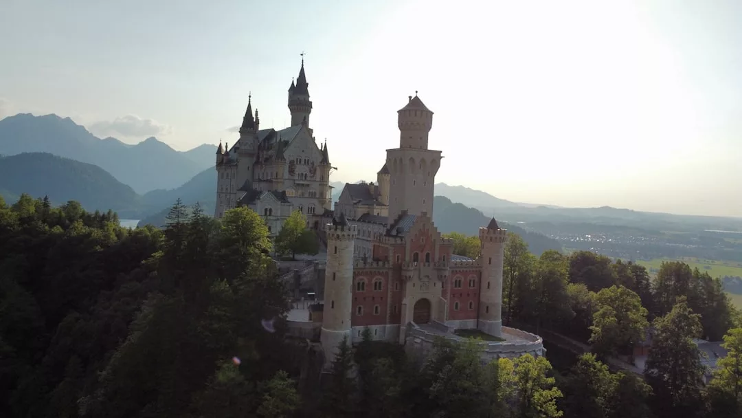 Auf einem bewaldeten Hügel auf Mallorca, umgeben von Bergen und einem Tal unter einem strahlenden Himmel, thront eine große Burg mit hohen Türmen.